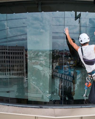 Industrial mountaineering worker hanging on rope and cleaning window of high-rise building while colleague standing behind glass. Man cleaner using safety equipment while washing skyscraper facade.